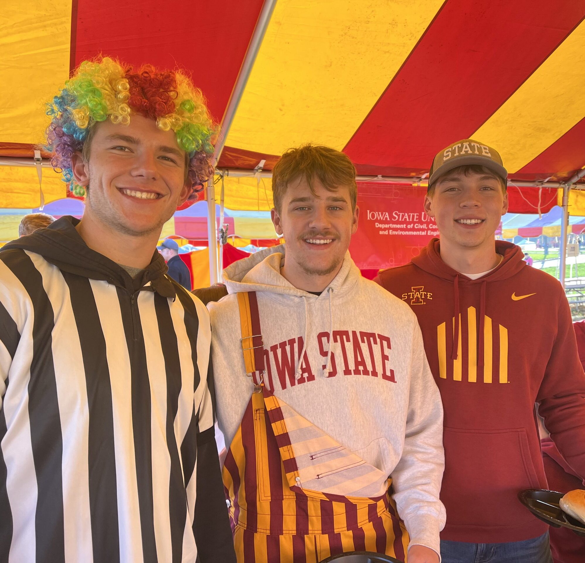 Three students stand together under a cardinal and gold striped tent and smile for the camera wearing Iowa State-themed clothing. The student on the left is wearing a rainbow wig.
