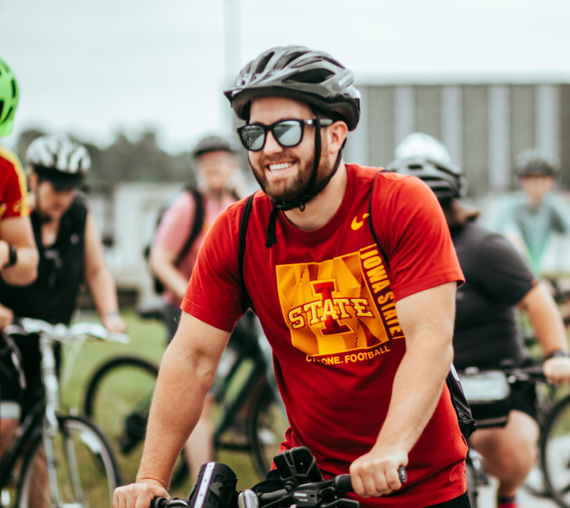 Man smiling in an Iowa State T-shirt on a bike with other bicycle riders socializing behind him.