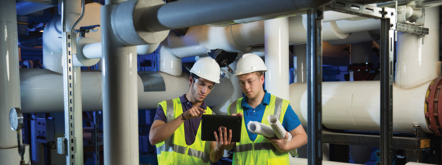 Students in hard hats and vests collaborating on a project with a pipe system in the background.