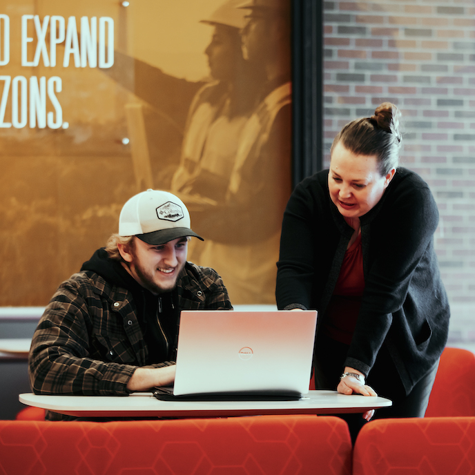 An advisor assists a student while he is on his laptop and they are conversing.