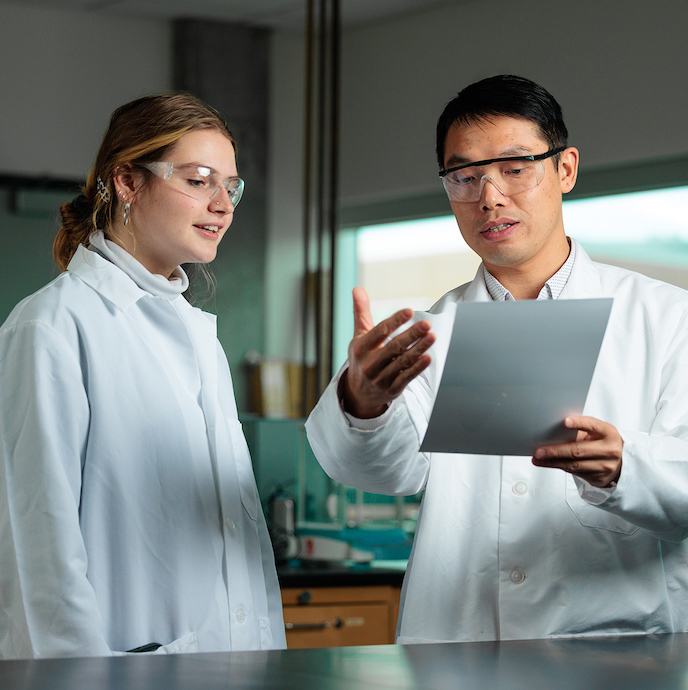 Students in qhite lab coats and safety goggles converse over a sheet of paper in a lab.